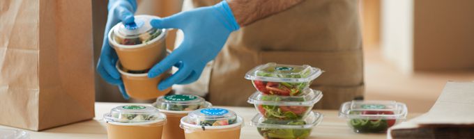 Man with blue rubber gloves loading clear plastic containers with salad and dressing into a paper bag